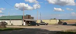 False-fronted building and sheet-metal building on paved street; farm fields in background