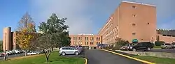 Wings of a three-to-four-story red brick building wrap around a parking lot filled with many cars.