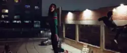 Dua Lipa and Miguel levitating over a brightly lighten parking lot, with a brick wall behind them.