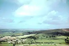 The houses and church of a small village can be seen in the bottom left of the picture. It is surrounded by a patchwork of fields with some trees on a hillside. Large hills in the distance.