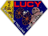 A diamond-shaped crest houses artworks of the Lucy fossil at left, the Lucy spacecraft at center, and an artist's impression of a Jupiter trojan. The word "Lucy" is written in a large, bold red font at the top right corner, while the words "First to the Trojans" and "SWRI · NASA · LM" are written in a smaller white font across the bottom edges of the diamond-shaped crest.