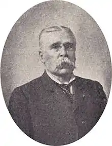 Head and shoulders of an older white man with a large drooping mustache wearing pince-nez glasses and a suit and tie. The portrait is surrounded by an oval frame and a depiction of an American flag.