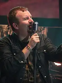 A singer, clothed in a shirt and a dark jacket, is holding a microphone and singing