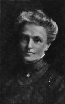 An older white woman, her white hair in a bouffant updo. She is wearing a high-collared dark dress or blouse.