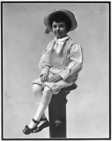 A black-and-white photographed of a neatly-dressed young boy in a hat, sitting on a pedestal