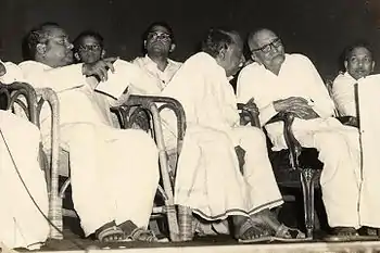  five men and a boy sitting in chairs. Four of the men are middle aged and one is in his seventies. One of the middle aged men is leaning toward and speaking to the old man.