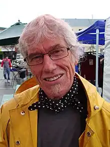 Portrait photo of a man in his late 60s with grey hair and glasses in a raincoat