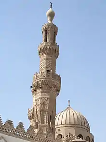 An ornate carved stone minaret, with a carved stone railing around three balconies, the first below its center, the second two thirds the way up, and the third near its top. The tip of the minaret is a large bulb-shaped stone decoration with a small bulb-shaped metal finial. Behind the minaret most of a dome is visible.