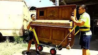 Removalists moving an upright piano with the a trolley and heavy straps connected to lifting harnesses