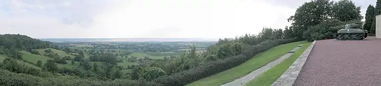 In the foreground tree-covered hills rising to the left and right frame a view over a valley. A tank and the edge of a building are on the rightmost hill. A flat plain of fields, trees and hedgerows fills the background, with a small hamlet visible in the middle-distance.