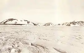 A mountain with a rounded summit and snowy slopes, with smaller hills to its right, rising from flat snow-covered ground