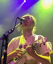 A singer with a ukulele in a white shirt and with glasses standing at a microphone at a concert, in a sea of multicoloured stage lighting