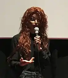 Narcissister wearing a mask and gesturing as she speaks.  She is standing in front of a projection screen.