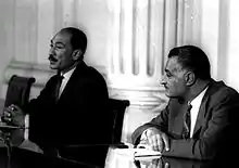 Two men in suits seated next to each other with their arms resting on a table