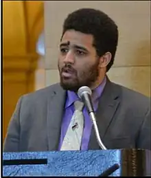 This is a close up portrait photo of Disability Advocate Noah McCourt. he is speaking at disability day on the hill and standing in the rotunda of the Minnesota State Capitol at a podium. He is surrounded by Marble walls