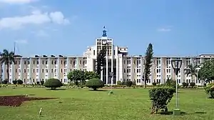 A wide four storied building with landscaped lawn and garden in the foreground