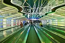 Photograph of a long tunnel illuminated by neon lighting in the ceiling and by glowing, colorful wall panels. The tunnel is about 10 meters (33 feet) wide, and there are four moving walkways in the center of the tunnel. There are several people on each walkway; far down the tunnel there are some staircases leading up.