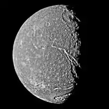 A round spherical body with its left half illuminated. The surface has a mottled appearance with bright patches among relatively dark terrain. The terminator is slightly to the right from the center and runs from the top to bottom. A large crater with a central pit can be seen at the terminator in the upper half of the image. Another bright crater can be seen at the bottom intersected by a canyon. The second large canyon runs from the darkness at the lower-right side to visible center of the body.