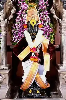 A stone icon of an arms-akimbo man standing on a brick and wearing a dhoti, angarkha (shirt), uparna (cloth across the chest), a crown and flower garlands till his waist.