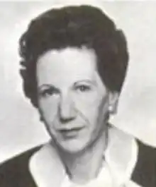 A middle-aged white woman with dark hair in a bouffant set, wearing a dark dress or jacket with a white collar