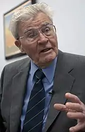 A grey-haired man wearing glasses, in a grey suit with blue shirt and tie.