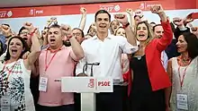 Pedro Sánchez wearing a white shirt and cheering with multiple people