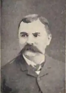 Head of a white man with a wide pointed mustache and neatly combed hair, wearing a dark suit coat and bow tie.