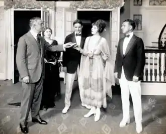 elegant elderly white man talking to vampish, extravagantly dressed young woman while middle-aged woman and two young men look on