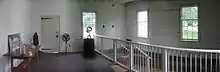  White pained interior of a room with wooden walls, three windows, and door. There is a wooden table at left and a white railing at left.