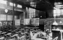 A black and white image of a large room with may chairs in front of a desk and charts. There are bars on the windows.