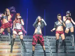 Five women standing in a row in front of a dark background. Two blonde, two brunette and a redhead. One of the brunettes is wearing a yellow top and a red wristband and holding a microphone to her mouth and singing into it. The other four are smiling. Two of them are wearing black tops and the other two are wearing white tops.