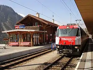 Two-story building with gabled roof next to double-track railway line and side platforms; a red train is on the near track