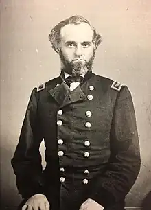 Sepia toned photo shows a man with a salt-and-pepper beard. He wears a dark military uniform with two rows of buttons.