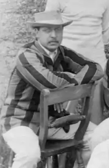 A black a white photograph of a man in a hat and stripped blazer sitting on a chair