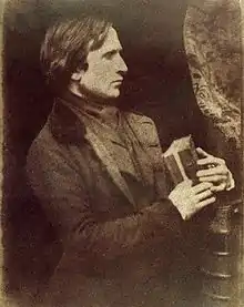 Calotype self-portrait photograph of Robert Adamson. He is standing sideways onto the camera, in profile, wearing a suit and holding a book in his hands.