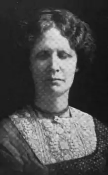 Portrait of a white woman against a dark background. Her eyes are closed. Her hair is wavy, parted at center, and dressed back. The yoke of her dress is heavily embroidered.