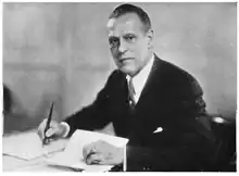 A photographic portrait of a man wearing a suit seated at a desk with a pen in his hand