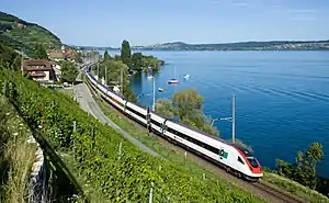 Red, white, and black trains pass a blue lake at speed