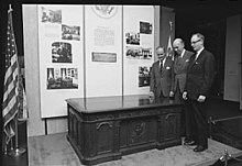  black and white image of three men in suits standing next to the Resolute desk. Museum text is on the wall behind them.