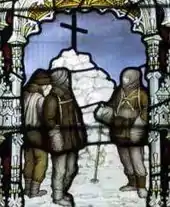 Three figures are depicted in coloured glass, standing by a cairn of snow topped by a large cross. The scene is framed by a decorative arch.