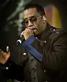 A black man is seen singing into a microphone while wearing a black-blue jacket and sunglasses. A multicolored LED screen is displayed in the background.