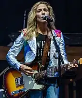 Sheryl Crow wearing a silver leather jacket, black shirt with white text, and blue jeans. She is standing in front of a microphone and singing while playing guitar