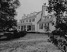 A large, two-story house with a veranda is surrounded by a lawn, manicured bushes and some trees.