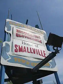 A white billboard with a blue ribbon around the edges. On the ribbon, "Cloverdale Town Centre" appear in yellow surrounding two banners. The top banner reads, "Destination Cloverdale" and the bottom reads, "Home of Smallville".