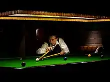 A man playing snooker on a baize-covered table, preparing to strike the white ball using a cue stick that he is holding with his right hand; the end of the cue stick is resting on the cross-shaped head of a long stick which he is holding with his left hand