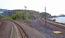 Two railroad tracks curving in opposite directions in a brushy area with water on the right and a high bridge on the left. The track on that side has cement ties and a third rail; the one on the right has wooden ties. It goes to a bridge in the background on that side, behind which is a wooded hill.