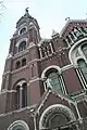 Ground-level view of a large, brick church with several stained glass windows, architectural niches, and a tall, tapering spire