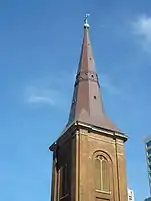 A detail of the tower and spire showing that it is octagonal and shaped rather like a witch's hat. In this view the copper has been renewed and appears brown and lustrous. On top are a cross and ball with green patina. The tower is brick, has flat corner pilasters and large louvred windows.