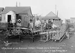 Ten men stand behind an array of empty dog sleds, some on racks. Most are Inuit and wear traditional or western working clothes: one is Caucasian and is dressed as a clergyman.