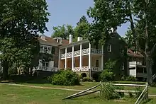 A large stone house with white dormers, window shutters and porch railings, surrounded by trees and shrubs.